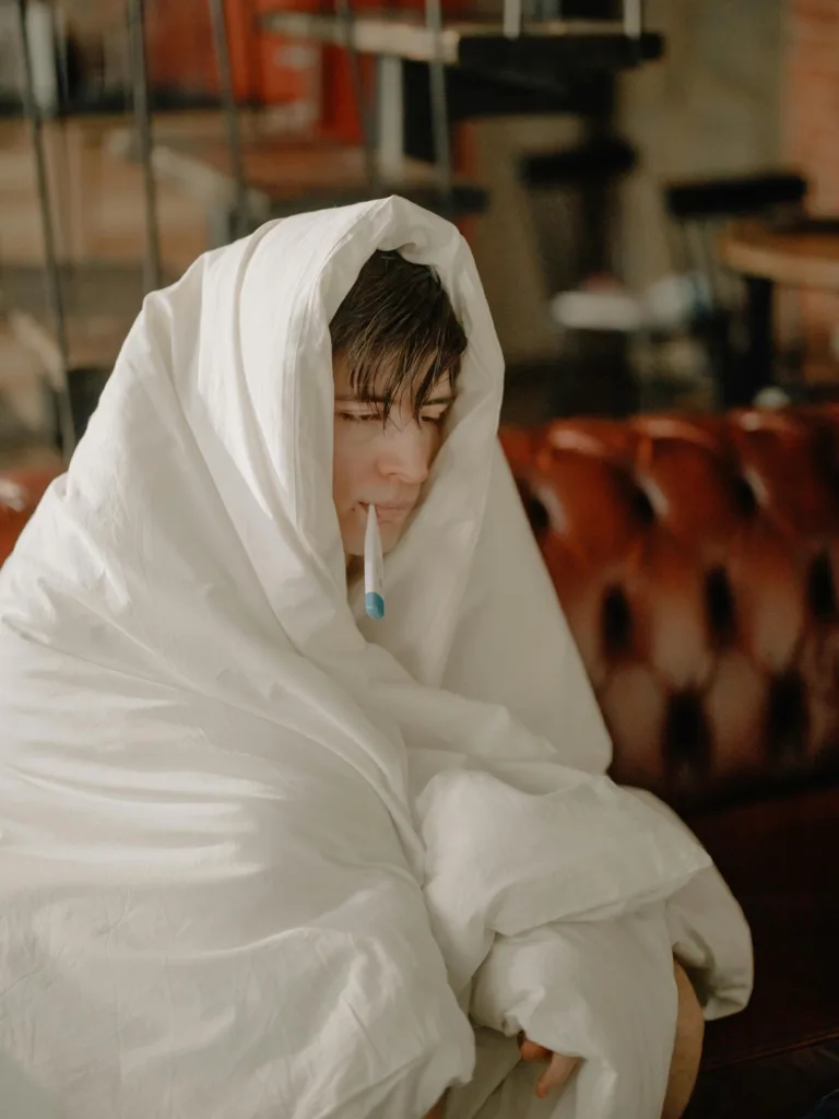 Person bundled in a blanket with a thermometer, indoors, feeling unwell.